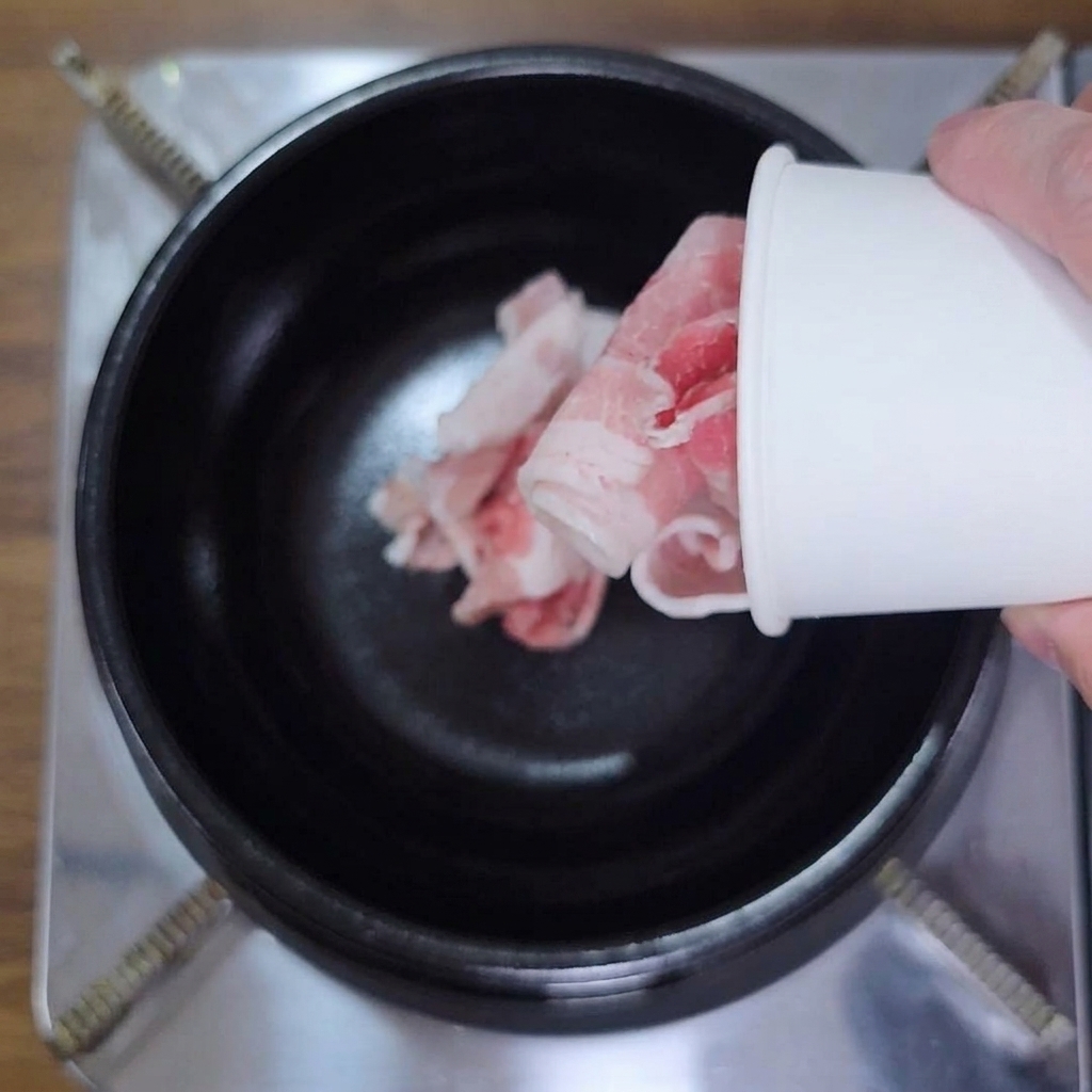 Mano vertiendo panceta cruda en láminas desde un recipiente de papel a una olla negra.