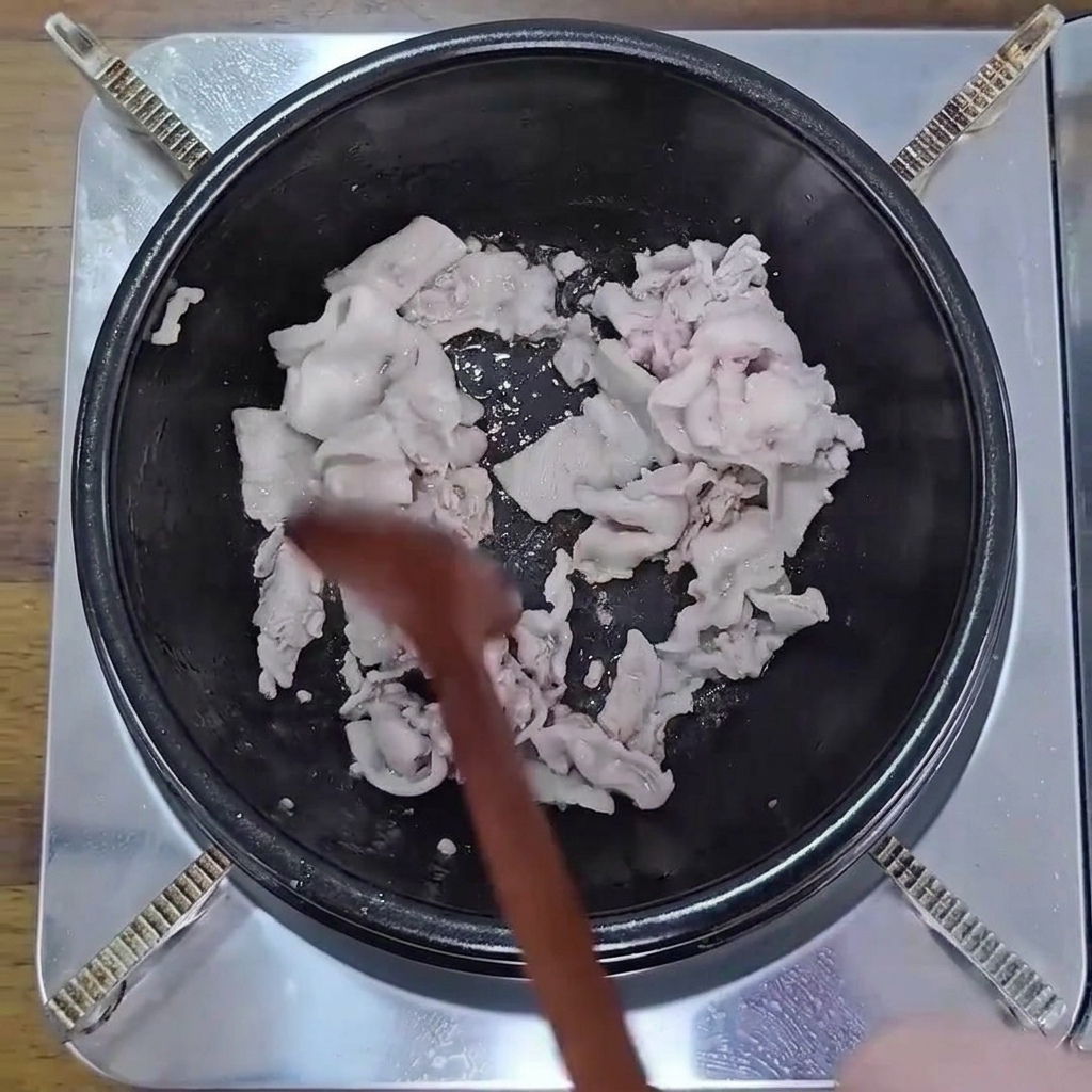 Láminas de panceta cocinándose en una olla negra con una cuchara de madera hasta dorarse ligeramente.