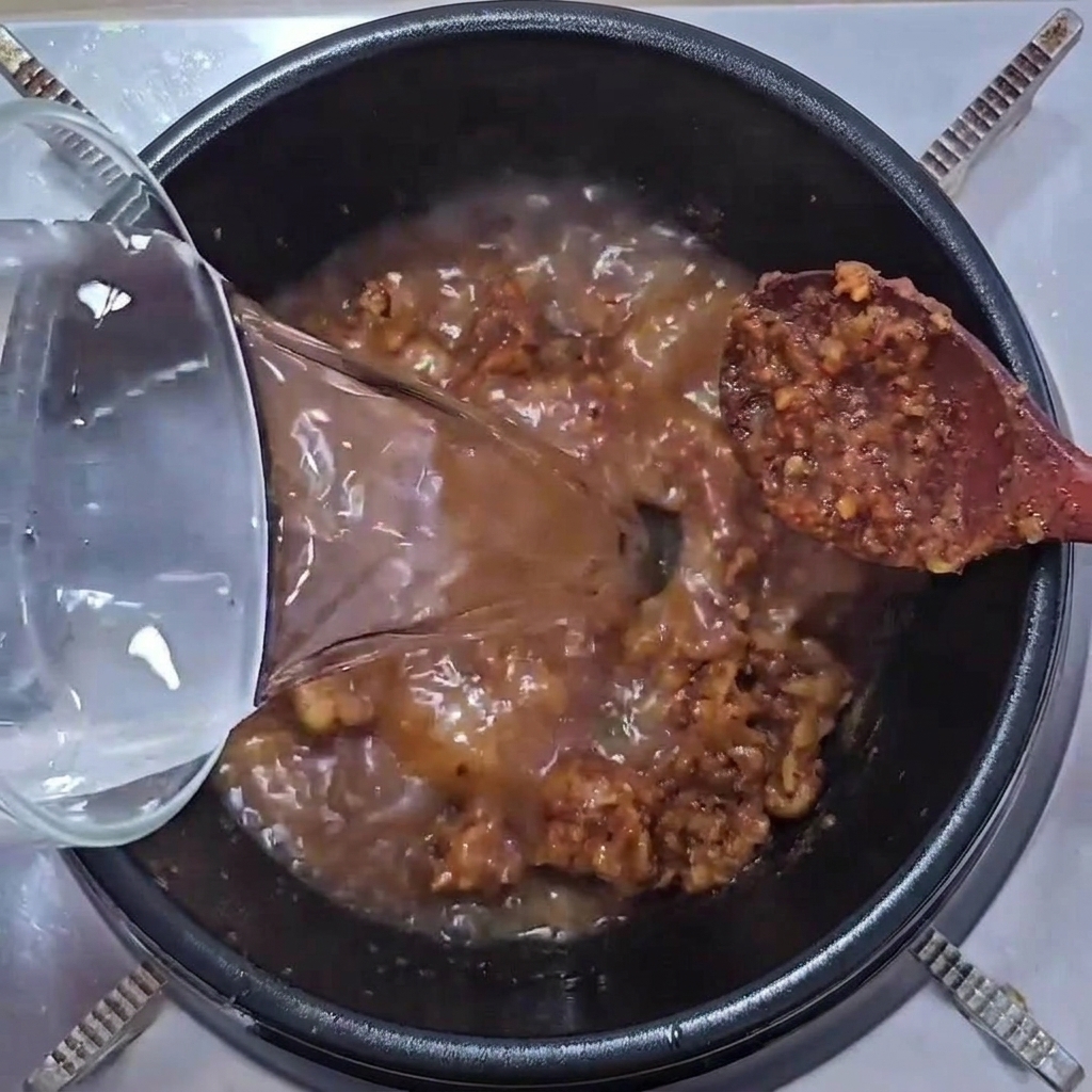 Agua vertiéndose en una olla con la mezcla chisporroteante de carne y pasta de soja.