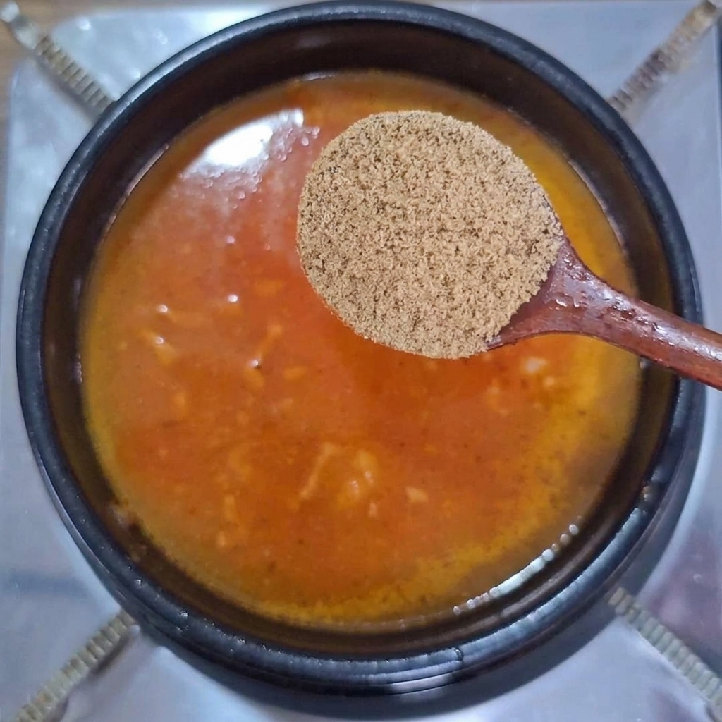 Cuchara de madera sosteniendo caldo de res en polvo sobre un caldo burbujeante de color rojo anaranjado.