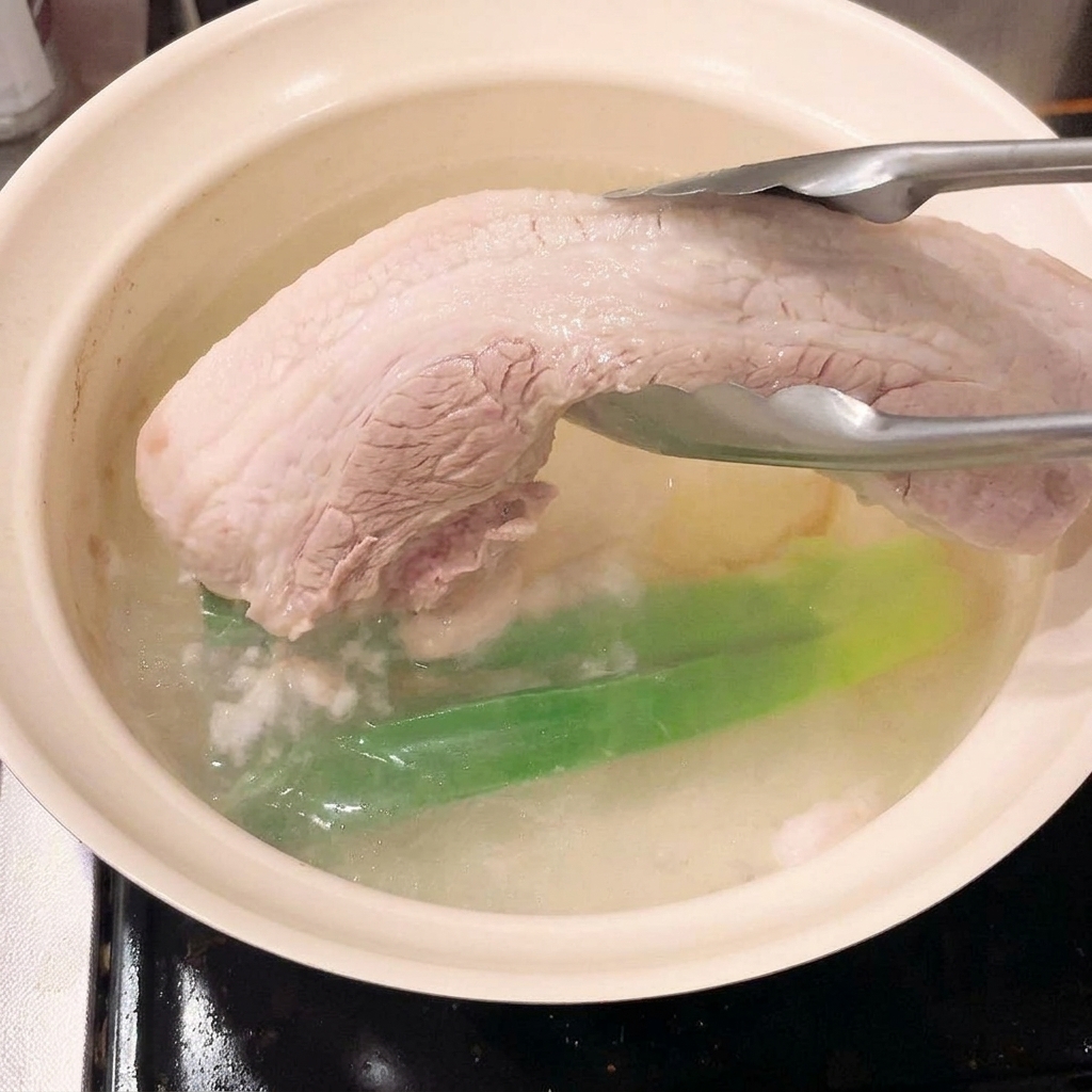 Pinzas levantando la pieza de panceta blanqueada y firme fuera del agua de cocción.