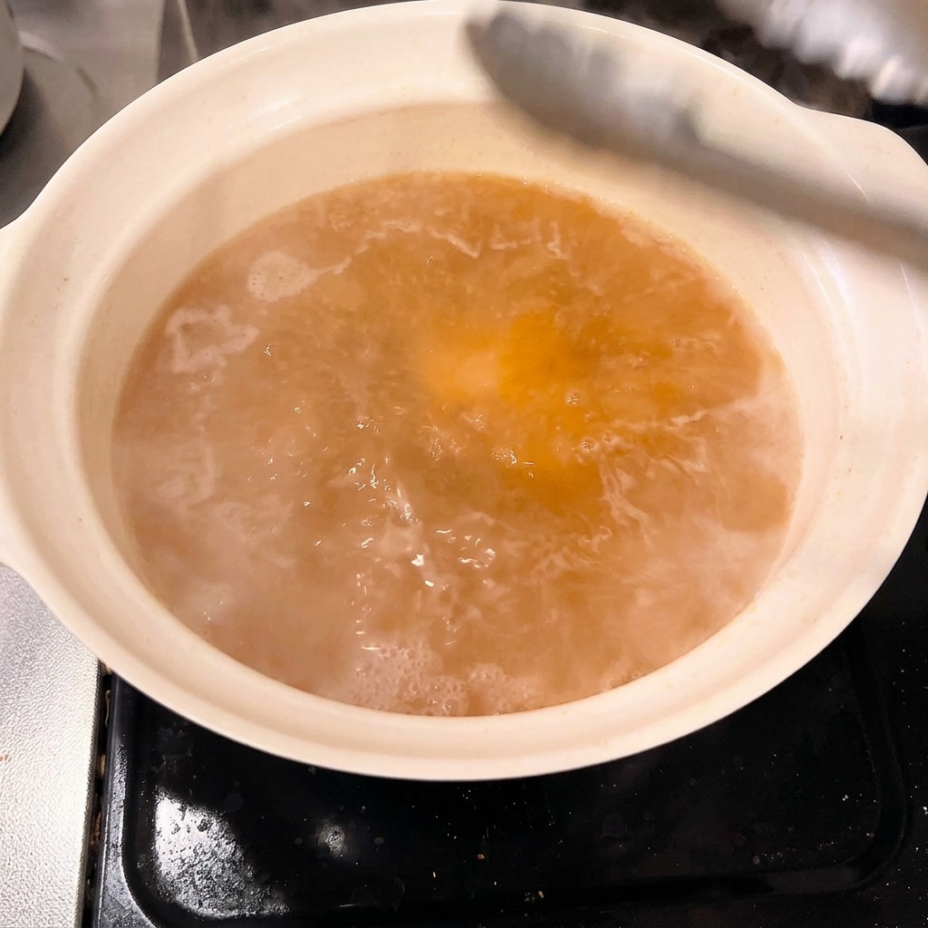 Trozos gruesos de panceta blanqueada siendo colocados cuidadosamente en la olla de caldo de miso con pinzas.