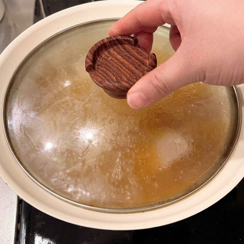 Una pequeña tapa de madera siendo colocada directamente sobre los trozos de panceta en el caldo.