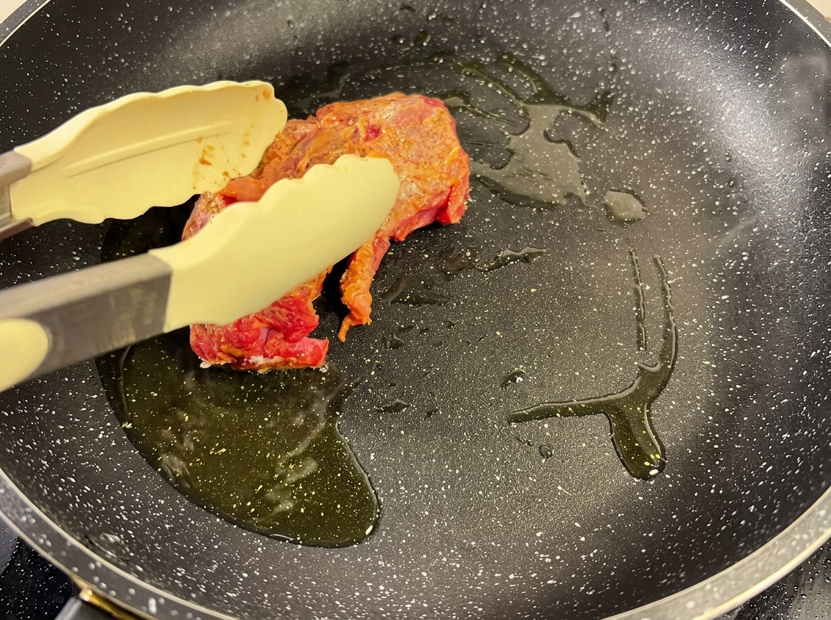 Un trozo de carne sazonada dorándose en una sartén caliente con aceite, sujetada con unas pinzas.