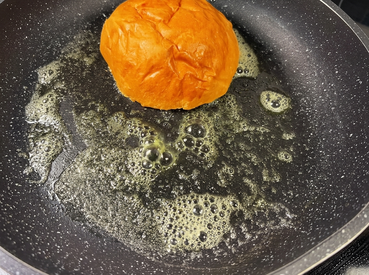 Un pan de hamburguesa tostándose en una sartén caliente con mantequilla derretida, adquiriendo un color dorado.