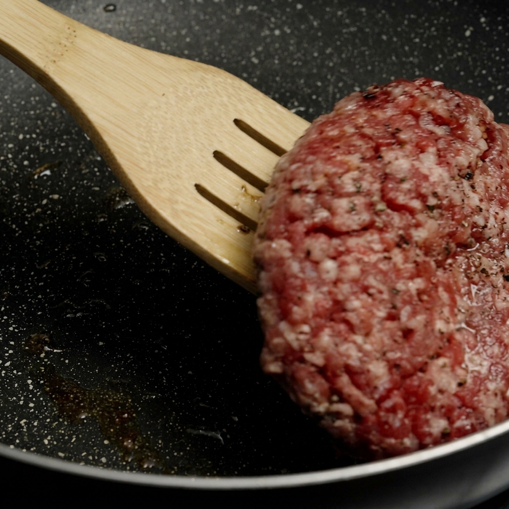 Una espátula de madera levantando y dando la vuelta a una hamburguesa gruesa con una base dorada y crujiente en una sartén caliente.