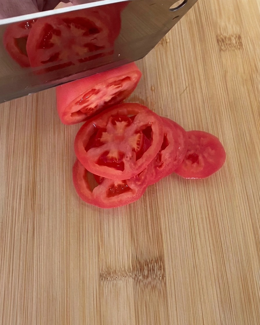Un cuchillo cortando un tomate rojo maduro en rodajas gruesas sobre una tabla de cortar de madera.
