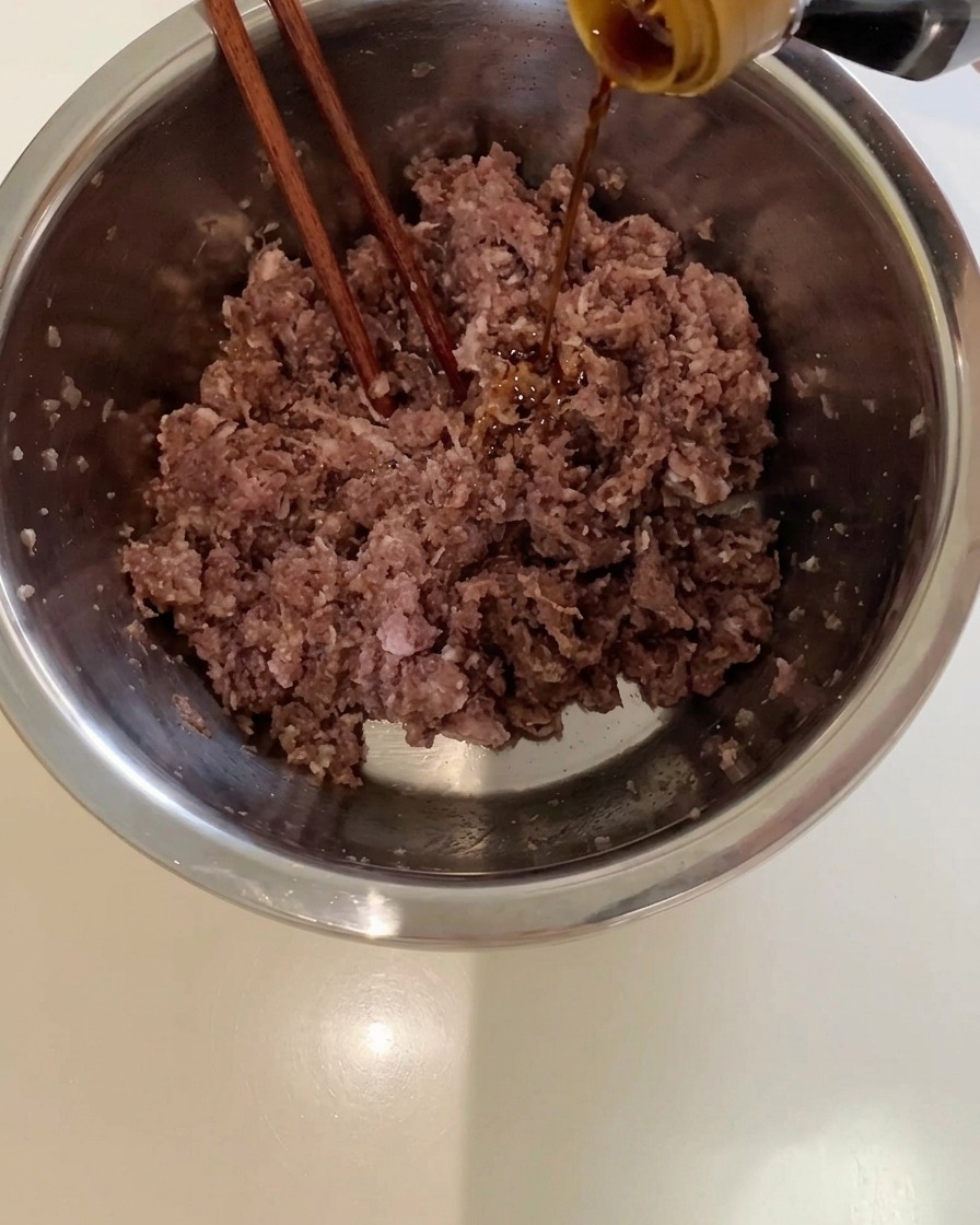 Salsa de soja oscura vertiéndose desde una botella en un bol de metal con carne picada.