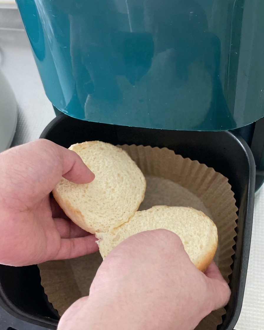 Dos mitades de un pan de hamburguesa blanco colocándose en la cesta de una freidora de aire forrada con papel pergamino.