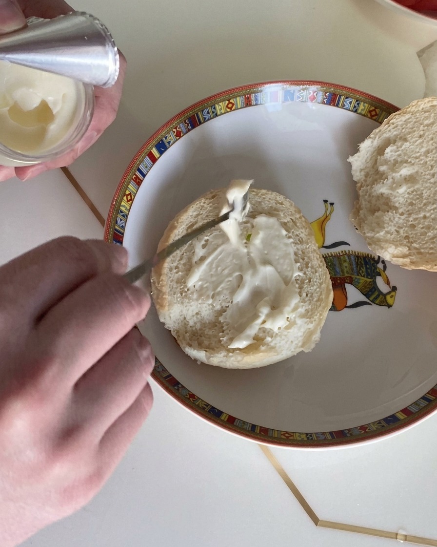 Primer plano de una cuchara untando un aderezo cremoso sobre la cara cortada de un pan de hamburguesa esponjoso.