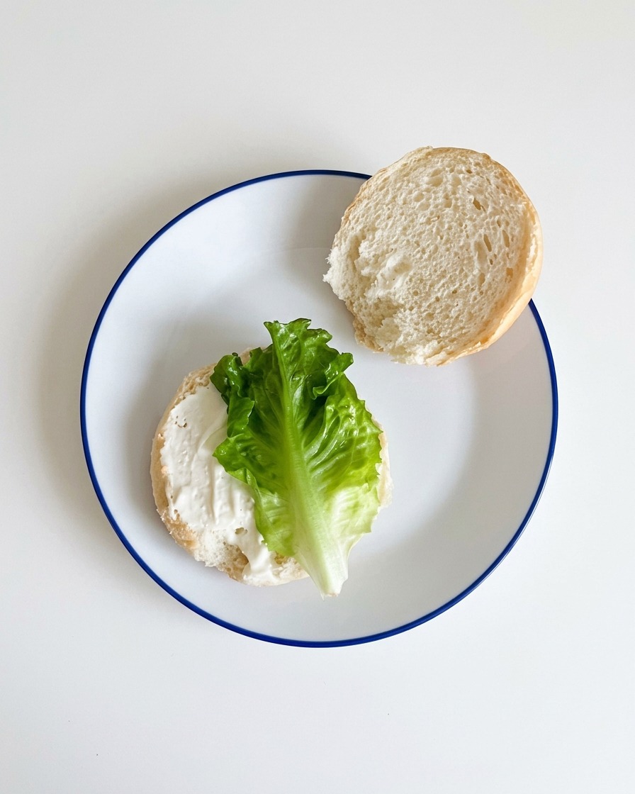Lechuga verde fresca colocándose sobre la mitad inferior de un pan de hamburguesa untado con salsa.