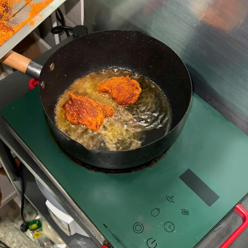 Trozos de carne de cerdo sazonados friéndose en aceite caliente dentro de una sartén negra sobre una estufa de inducción verde.