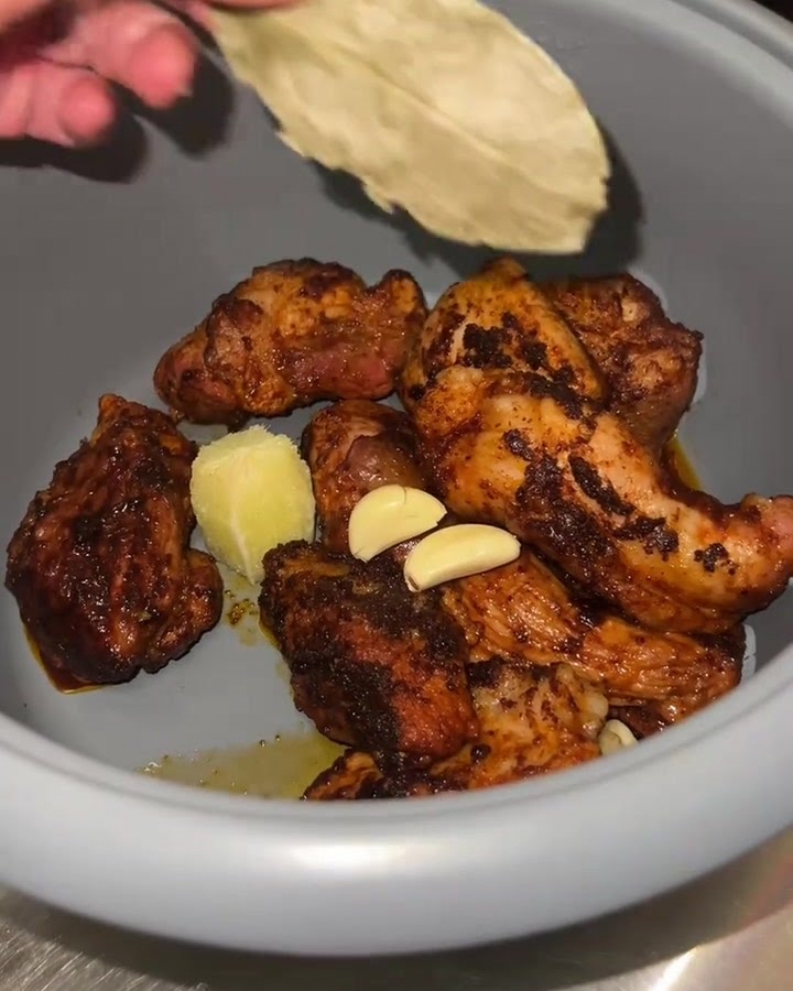 Primer plano de una olla arrocera con trozos de cerdo dorados, ajo fresco, pulpa de lima y una hoja de laurel siendo añadidos.