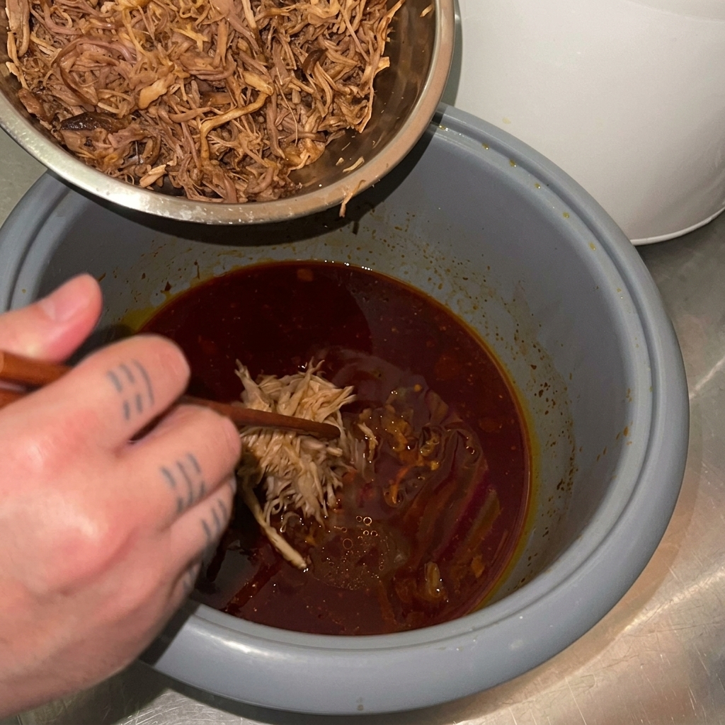 Cerdo desmechado siendo transferido desde un tazón de metal de nuevo a una olla arrocera que contiene una salsa de braseado de color rojo oscuro.