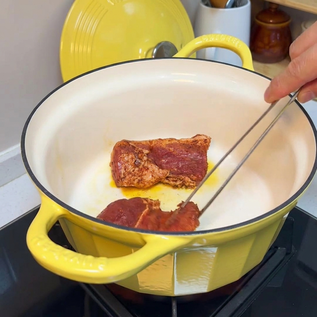 Pinzas girando trozos de cerdo sazonados en una olla de hierro fundido amarilla para sellar los bordes.