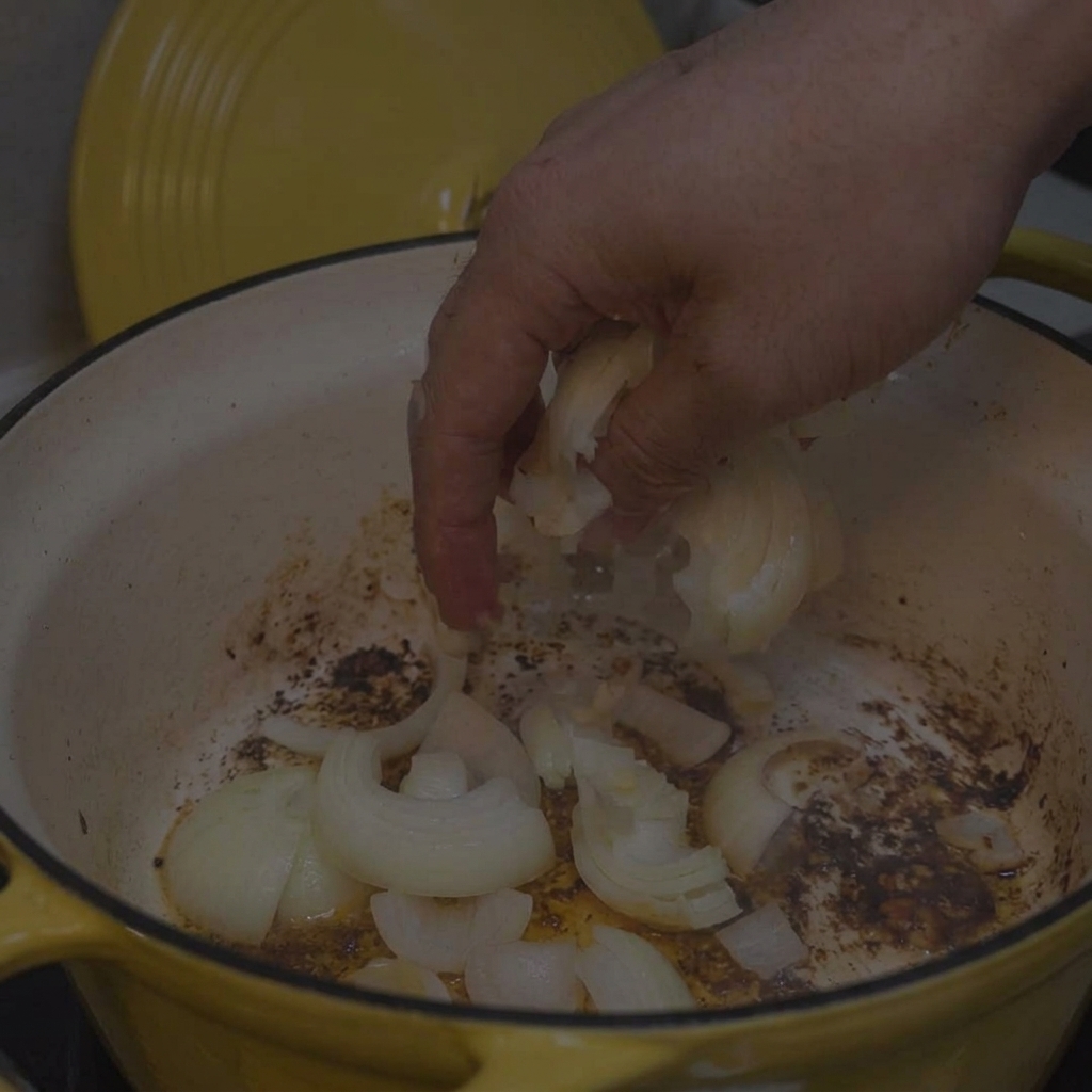 Una mano echando cebollas blancas en rodajas en una olla amarilla con restos dorados en el fondo.