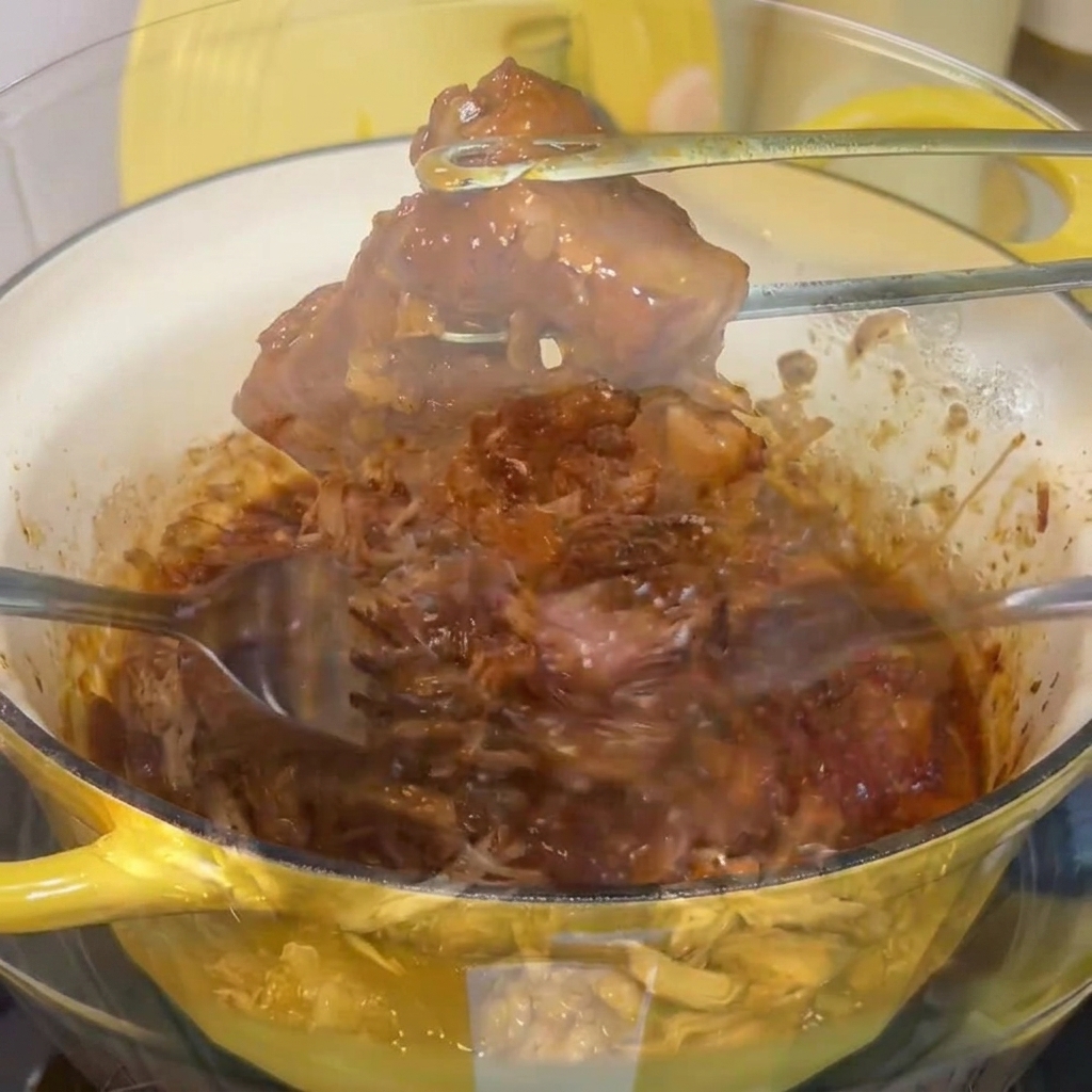 Carne de cerdo cocida y tierna siendo deshebrada con dos tenedores en un bol de mezcla de vidrio transparente.