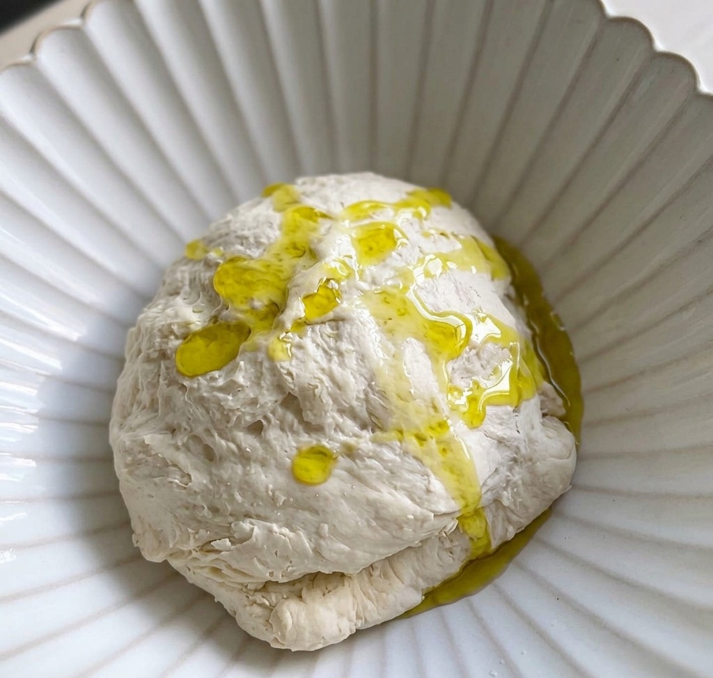 Un chorrito de aceite de oliva vertiéndose sobre una bola de masa en un recipiente blanco.