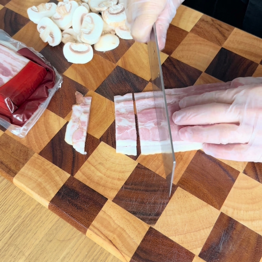 Manos con guantes usando un cuchillo de cocina para cortar tiras de tocino crudo en pequeños rectángulos sobre una tabla de madera a cuadros.