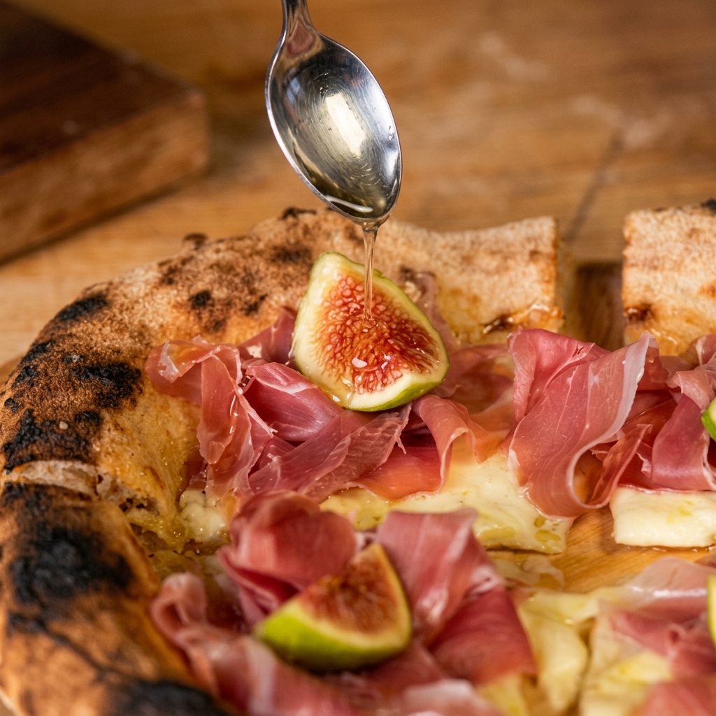 Cuchara de plata rociando miel clara sobre una pizza rústica con queso fundido, lonchas de jamón de Parma y medio higo fresco.