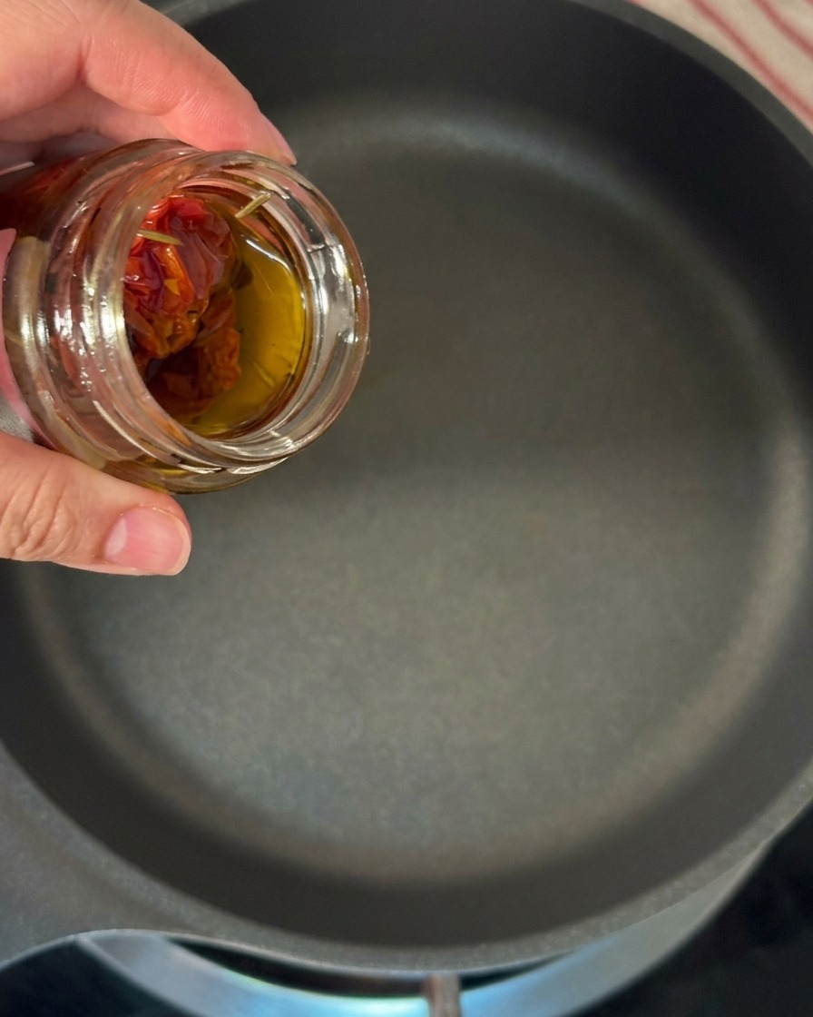 Una mano vertiendo el aceite infusionado de un frasco de tomates secos en una sartén grande.