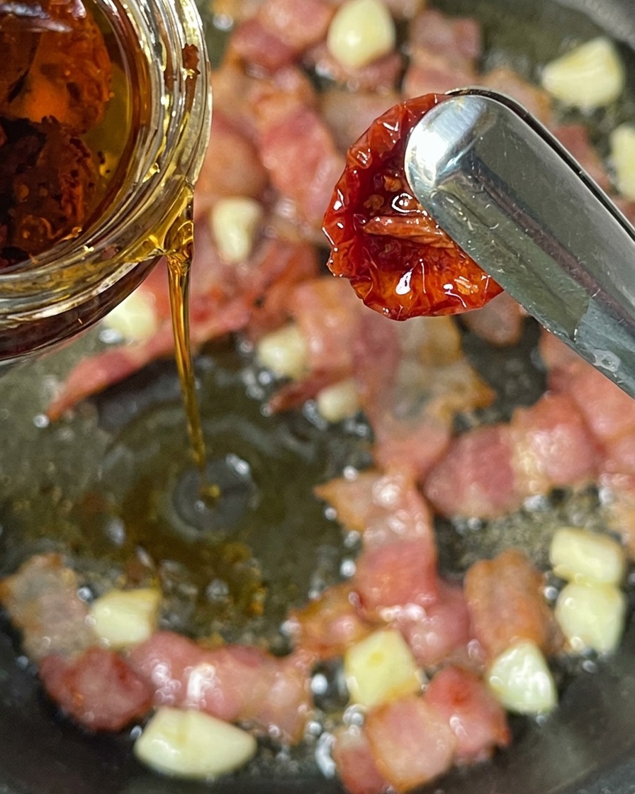 Tomates secos siendo añadidos a una sartén con tocino y ajo.