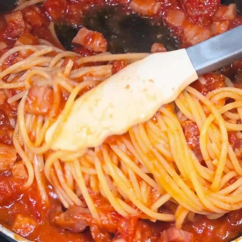 Espaguetis mezclándose con pinzas en la sartén, cubiertos de salsa roja brillante.