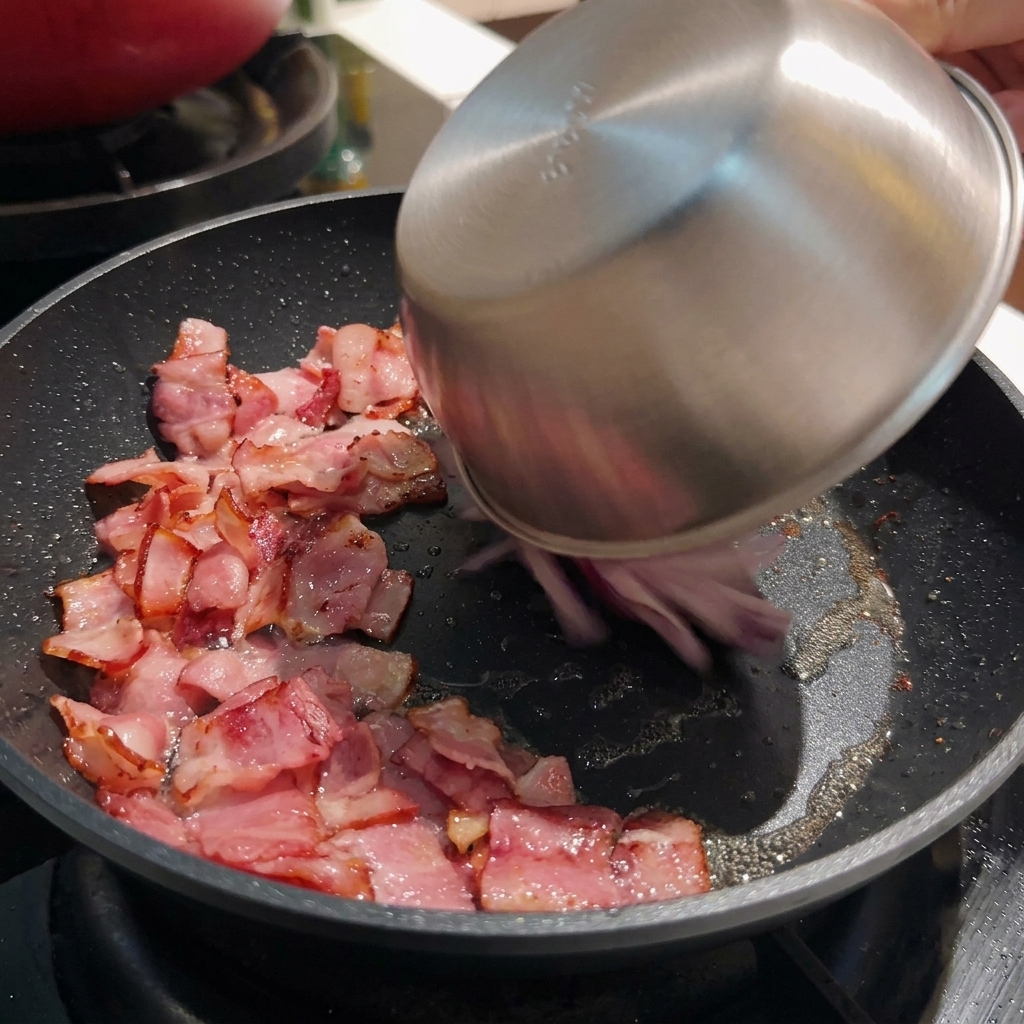 Cebollas moradas en rodajas siendo vertidas desde un tazón de acero inoxidable a una sartén caliente que contiene trozos de tocino crujiente.