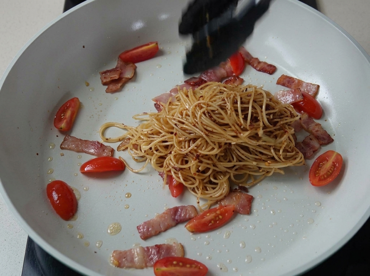Espaguetis sazonados mezclándose con bacon y tomates en una sartén.