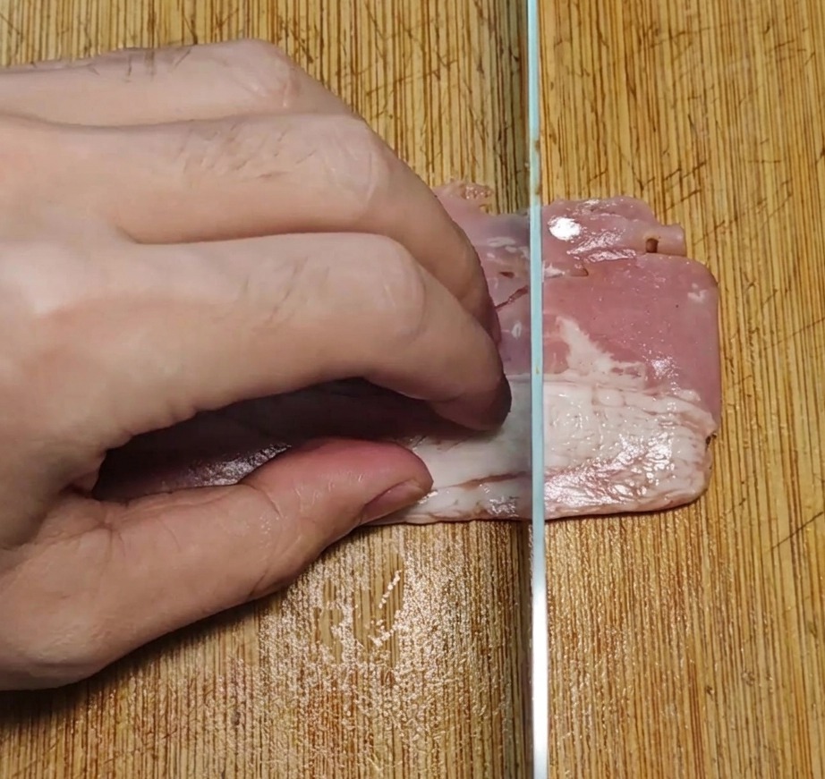 Tiras de bacon siendo cortadas en piezas rectangulares uniformes sobre una tabla de madera.