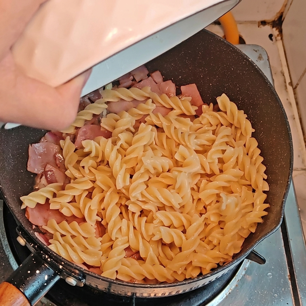 Pasta fusilli y trozos de espárragos siendo añadidos a la sartén con el bacon y la cebolla.