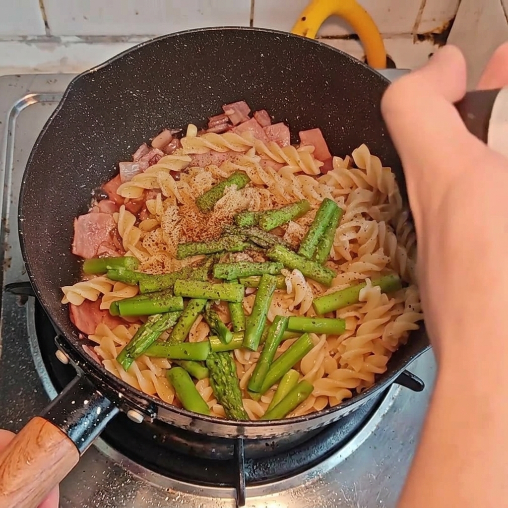 Diversos condimentos siendo espolvoreados y vertidos sobre la pasta, el bacon y los espárragos en la sartén.