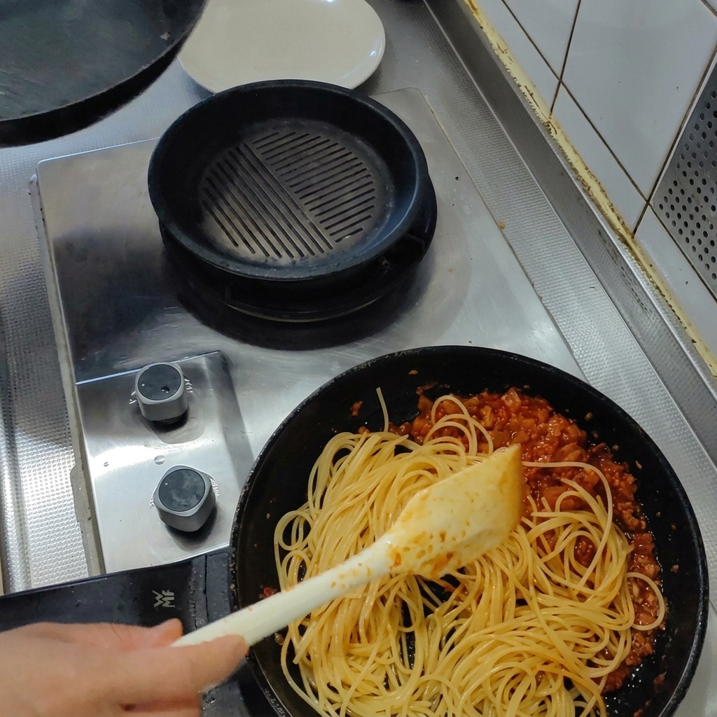 Espaguetis cocidos mezclándose en una sartén con salsa espesa de tomate y tocino usando una espátula.