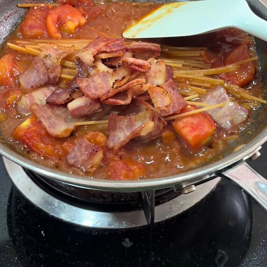 Trocitos de bacon frito esparcidos sobre la pasta y la salsa de tomate en una sartén.