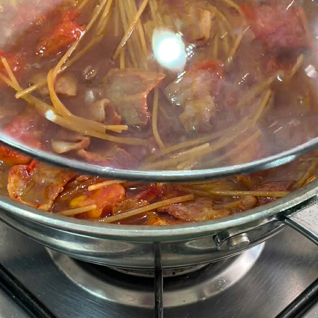 Espaguetis cociéndose en salsa de tomate bajo una tapa de cristal.
