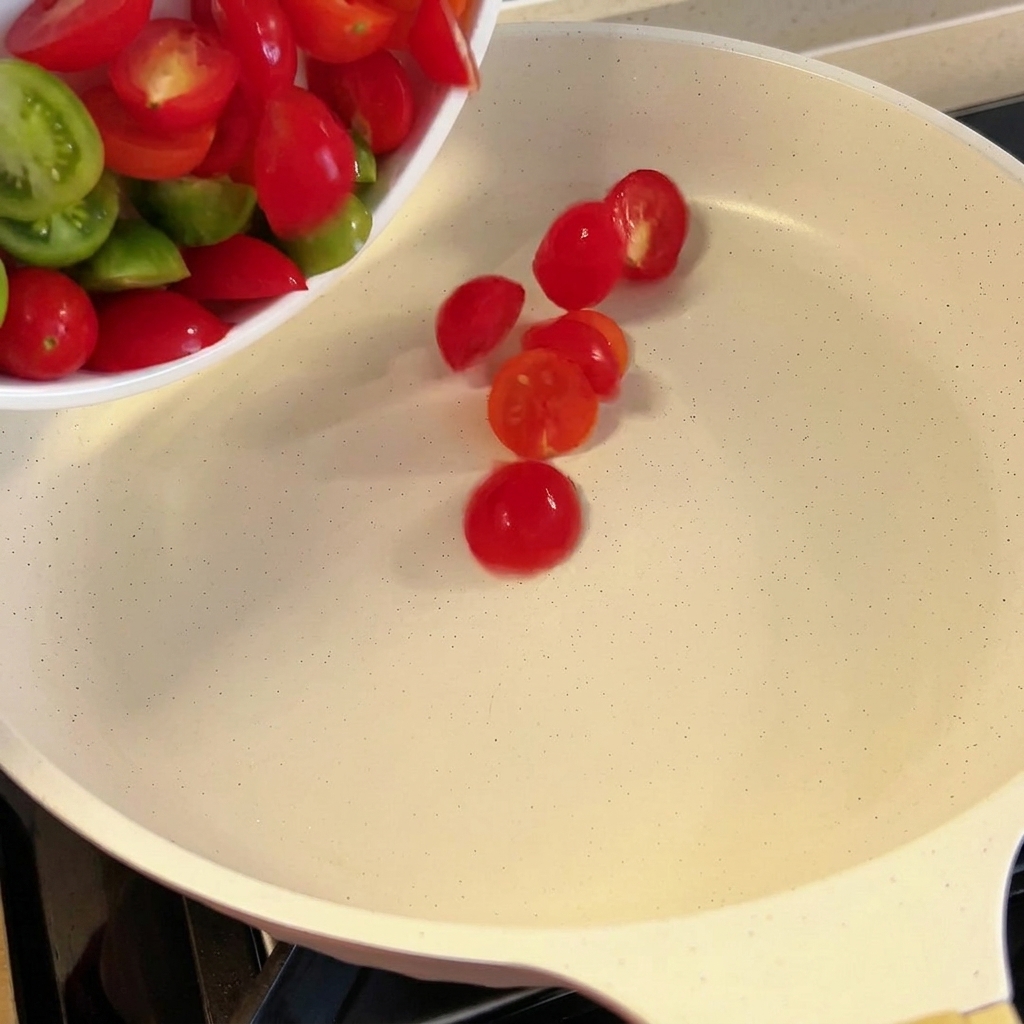 Tomates cherry frescos cortados por la mitad siendo añadidos a una sartén blanca.