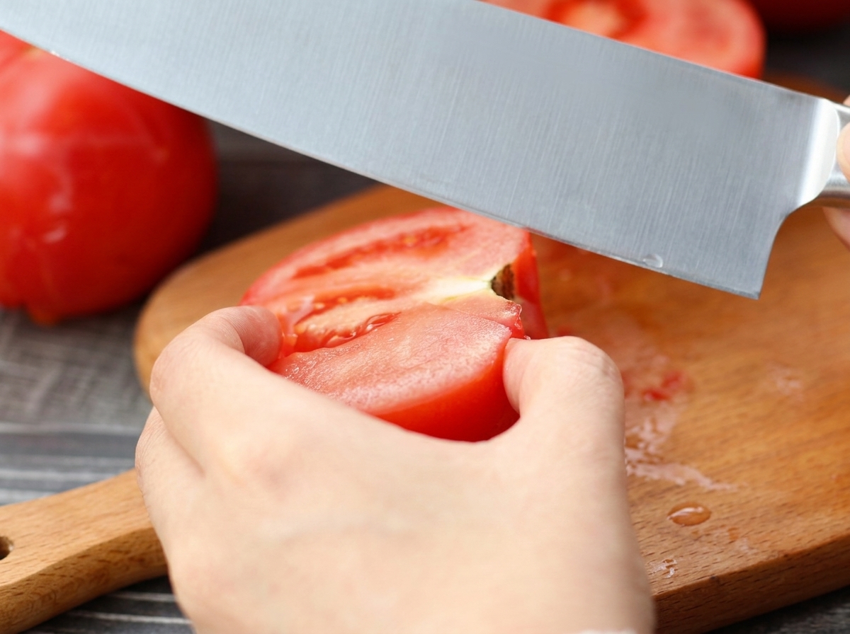 Un cuchillo rebanando un tomate pelado sobre una tabla de cortar de madera.