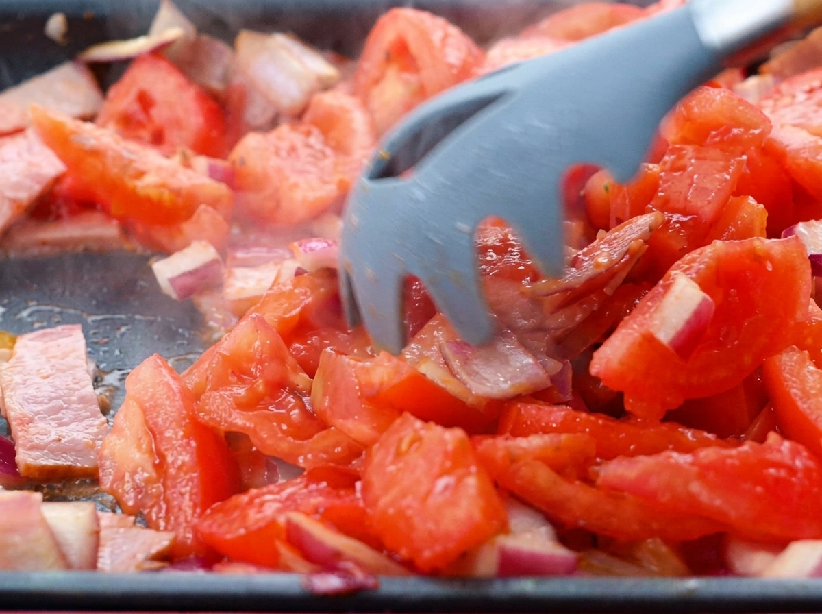 Tomates troceados incorporándose a una mezcla de tocino y cebolla en una sartén.