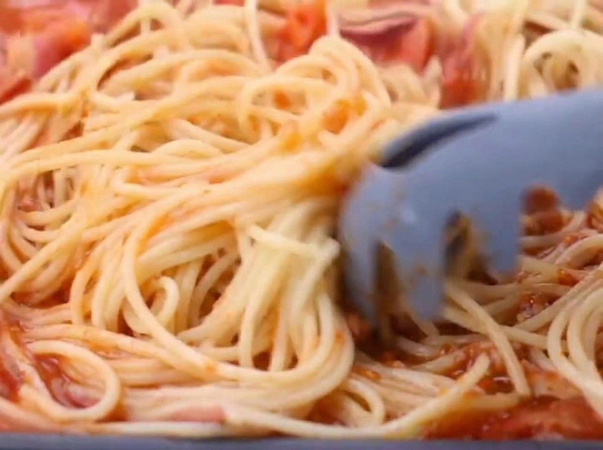 Espaguetis cocidos añadiéndose a la salsa de tomate y tocino.