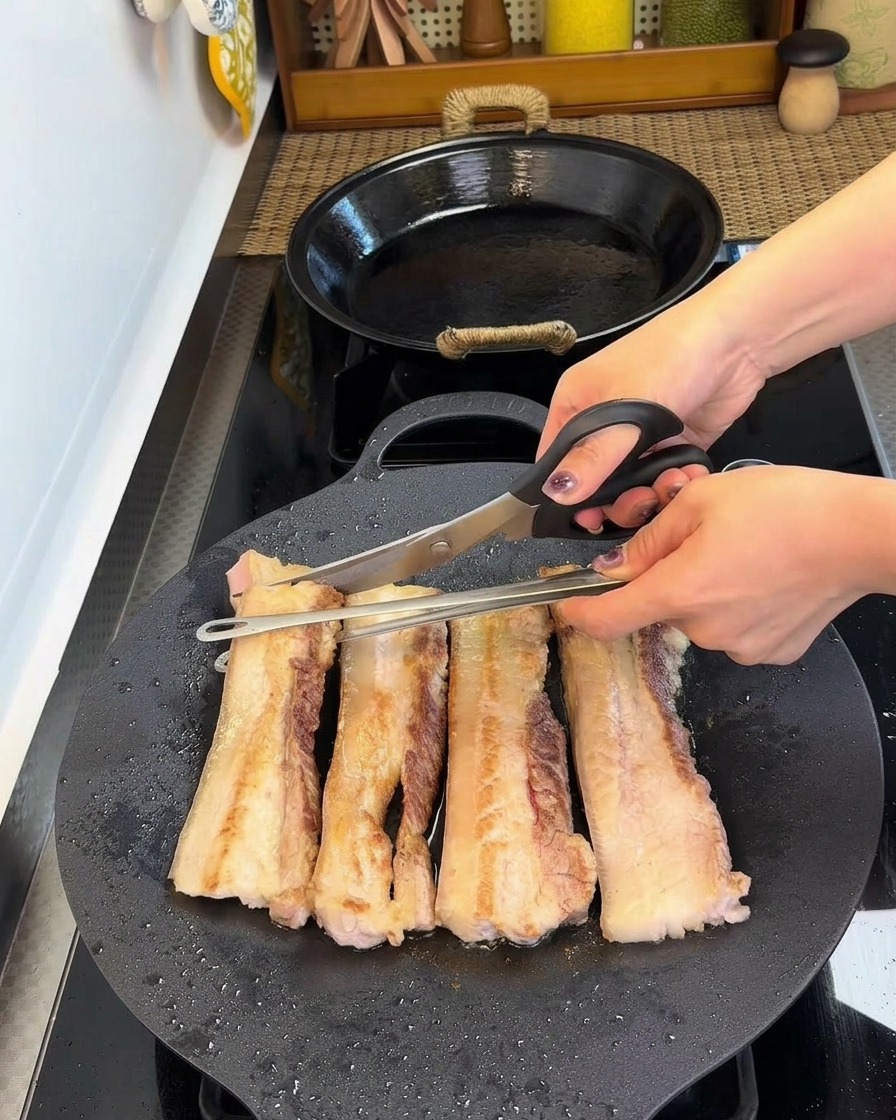 Tijeras de cocina cortando tiras de panceta dorada en trozos más pequeños dentro de la sartén.