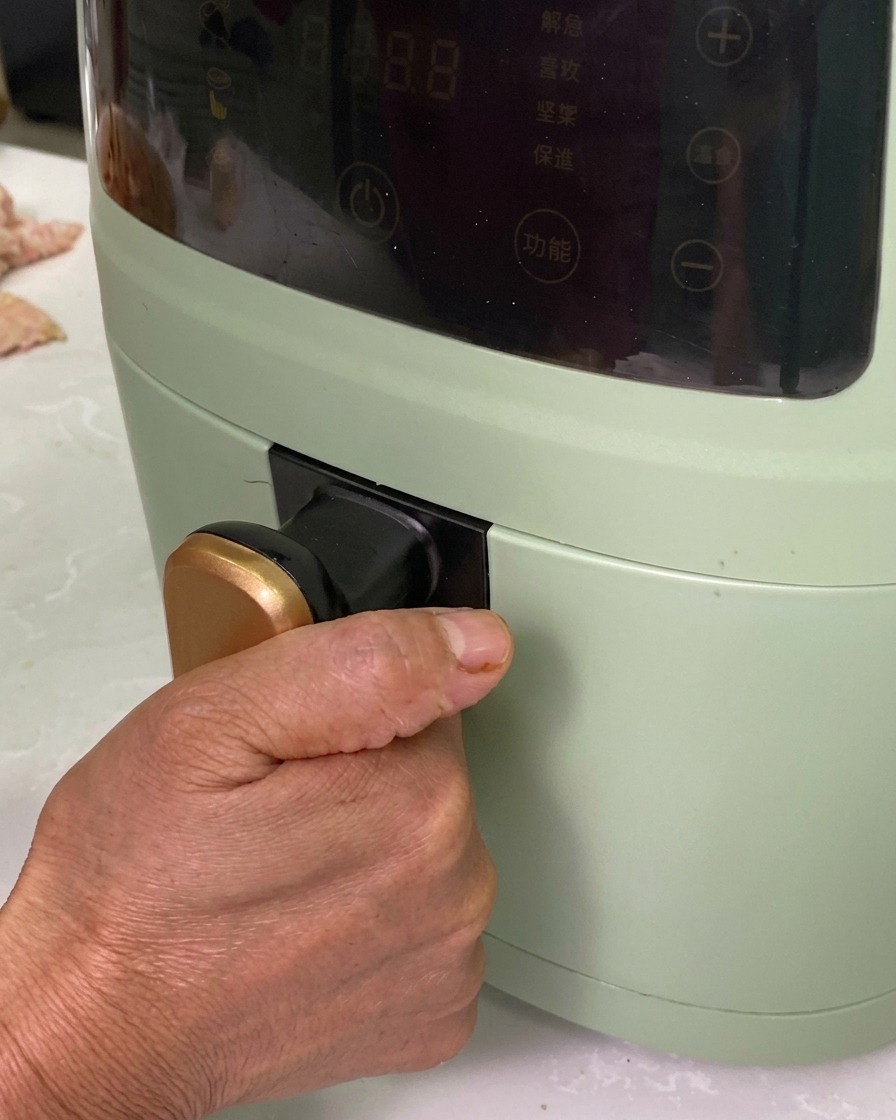 Una mano deslizando una bandeja de aluminio llena de rollitos sazonados en una freidora de aire verde.