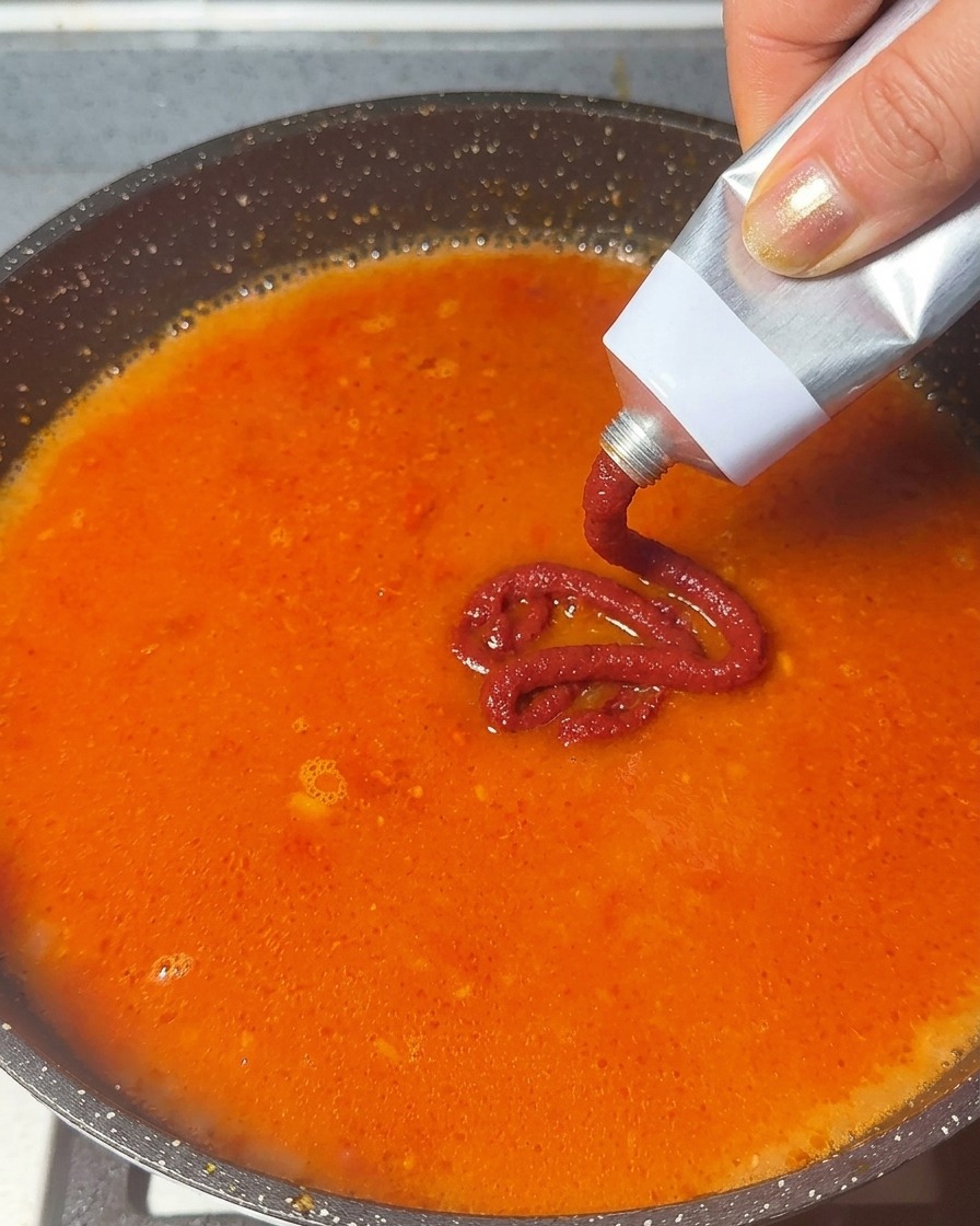 Pasta de tomate siendo exprimida desde un tubo de metal hacia una base de sopa roja anaranjada a fuego lento.