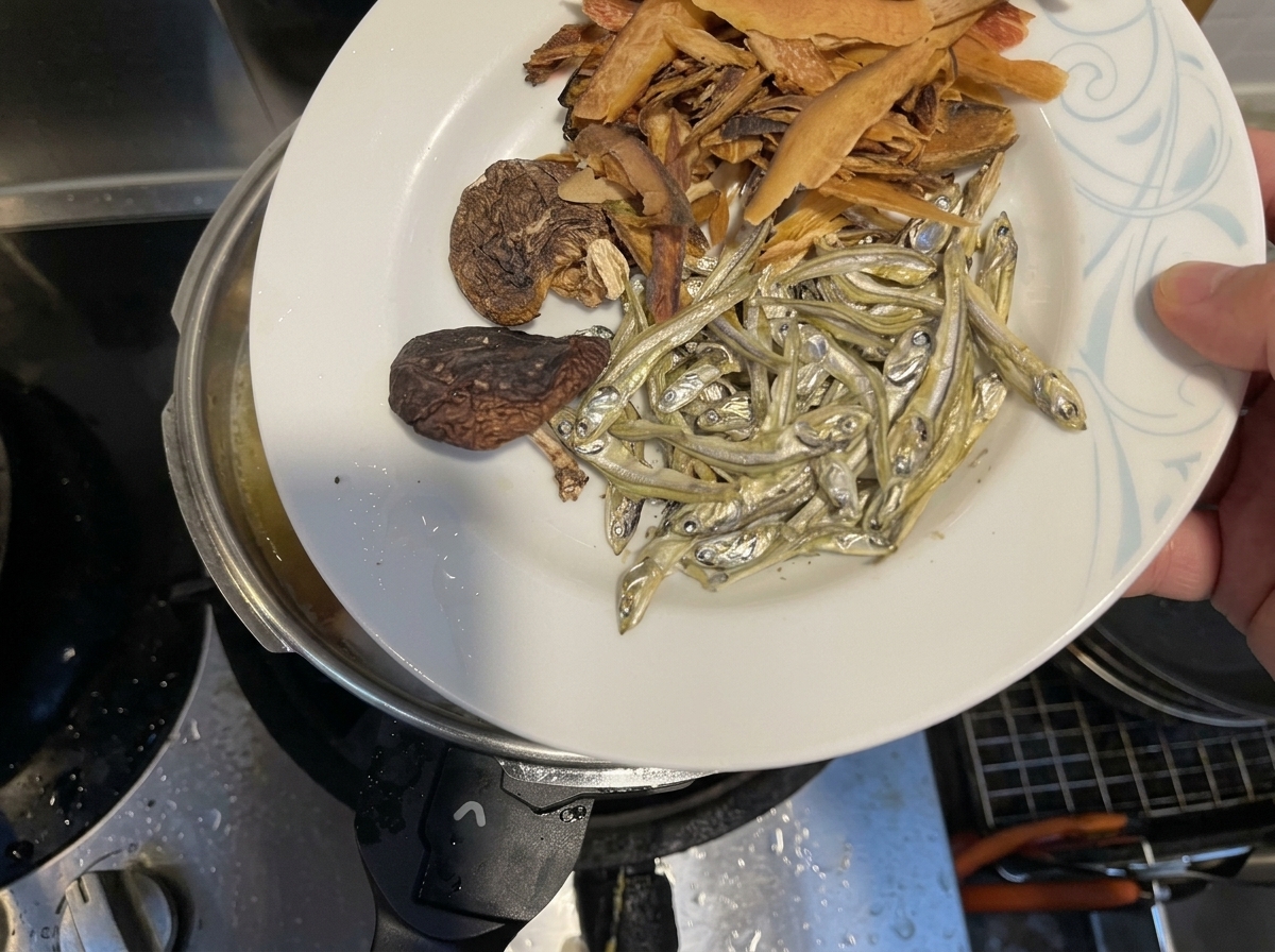 Pescado seco, setas shiitake y copos de pescado siendo añadidos a una olla de líquido hirviendo.