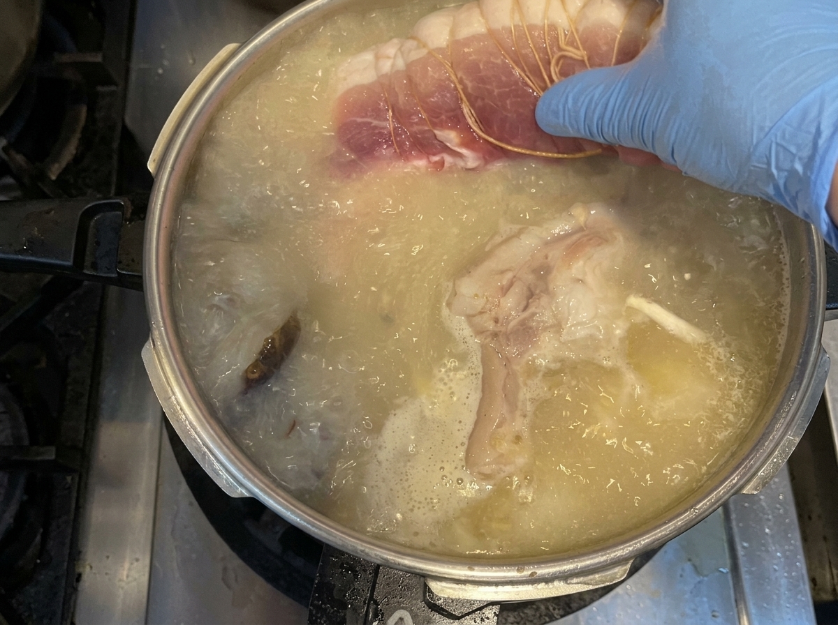 Una mano con un guante azul colocando un trozo de cabezada de cerdo cruda y atada en una olla con caldo y verduras.