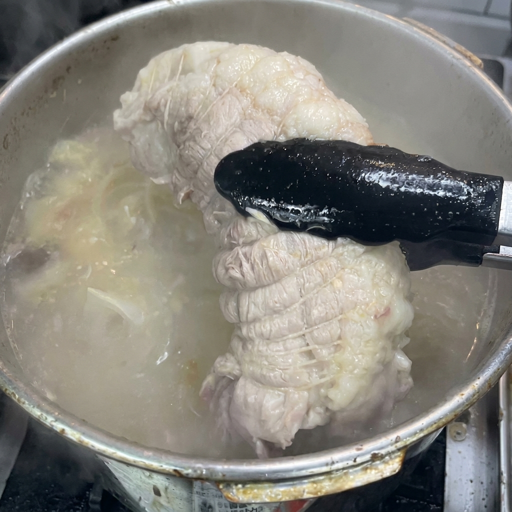 Pinzas negras levantando un trozo de cabezada de cerdo cocido y atado fuera de una olla burbujeante.
