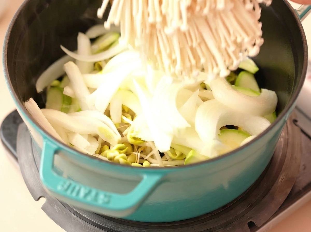Un racimo de setas enoki siendo colocado en una olla con cebolla, calabacín y brotes.