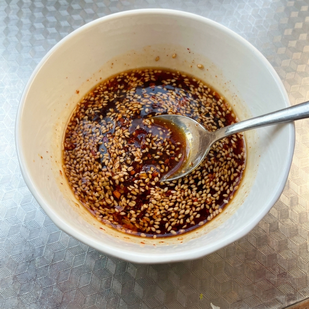 Primer plano de un cuenco blanco con una mezcla de salsa oscura y picante cubierta con semillas de sésamo, siendo mezclada con una cuchara.