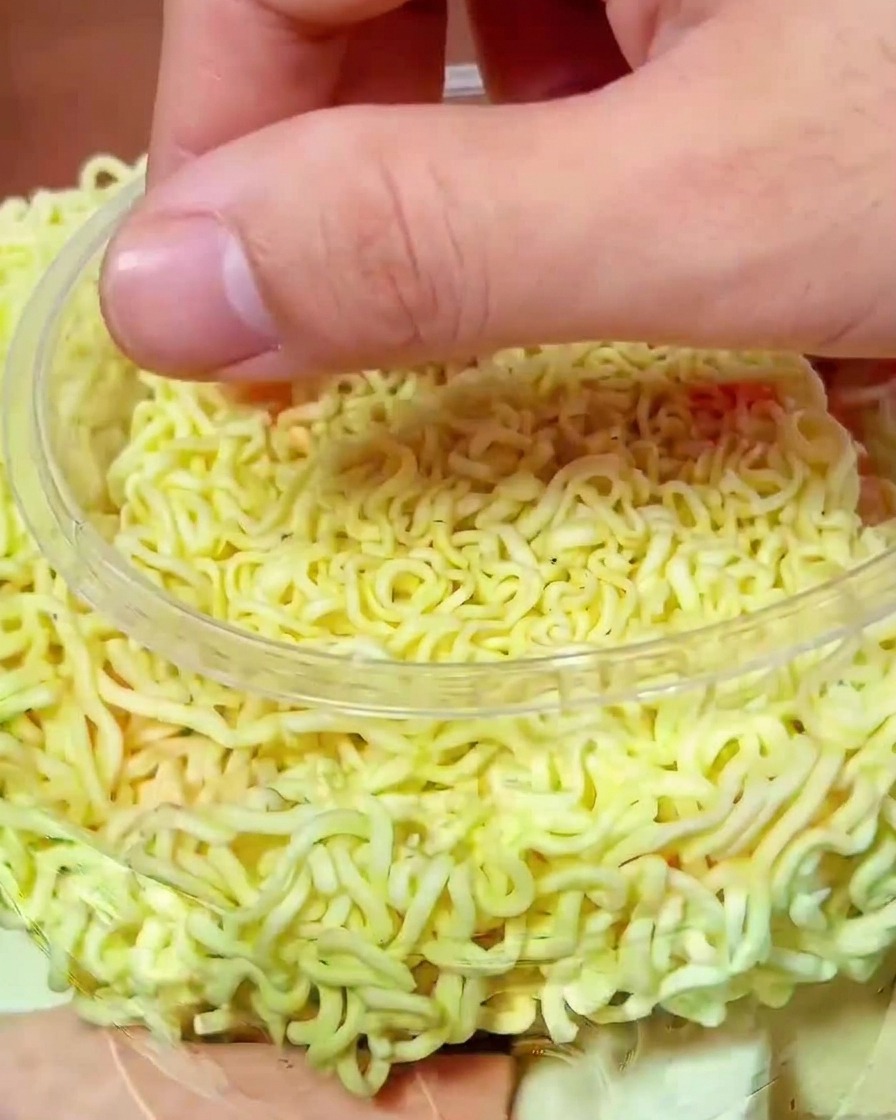 Una mano colocando trozos de kimchi rojo vibrante sobre los fideos crudos en la olla de cocción.