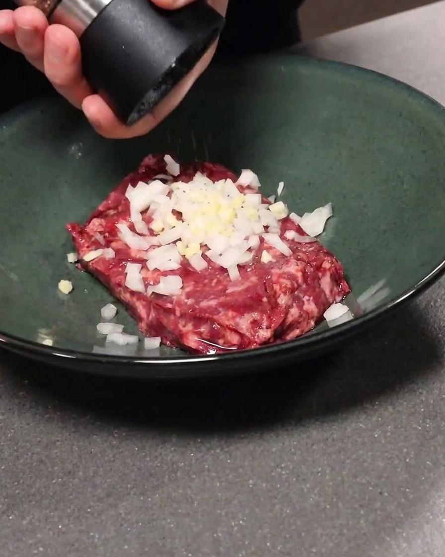 Carne picada mezclada con ajo y cebolla en un bol, mientras se le añade pimienta negra.