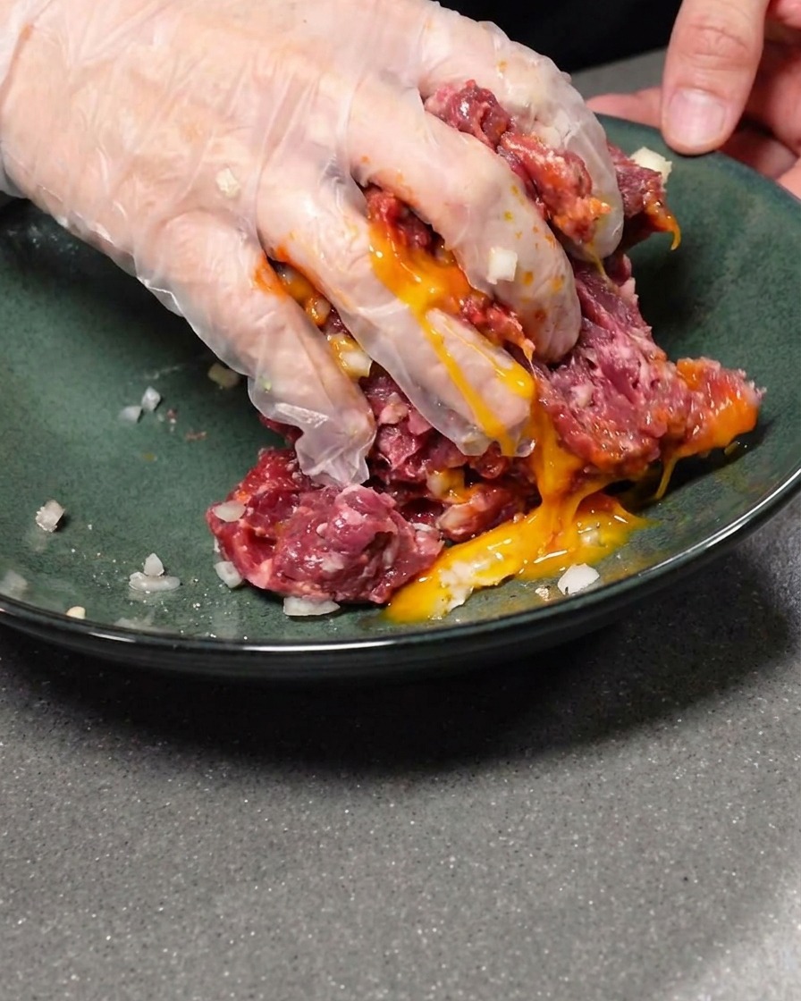 Una yema de huevo cruda incorporándose a la mezcla de carne molida sazonada.